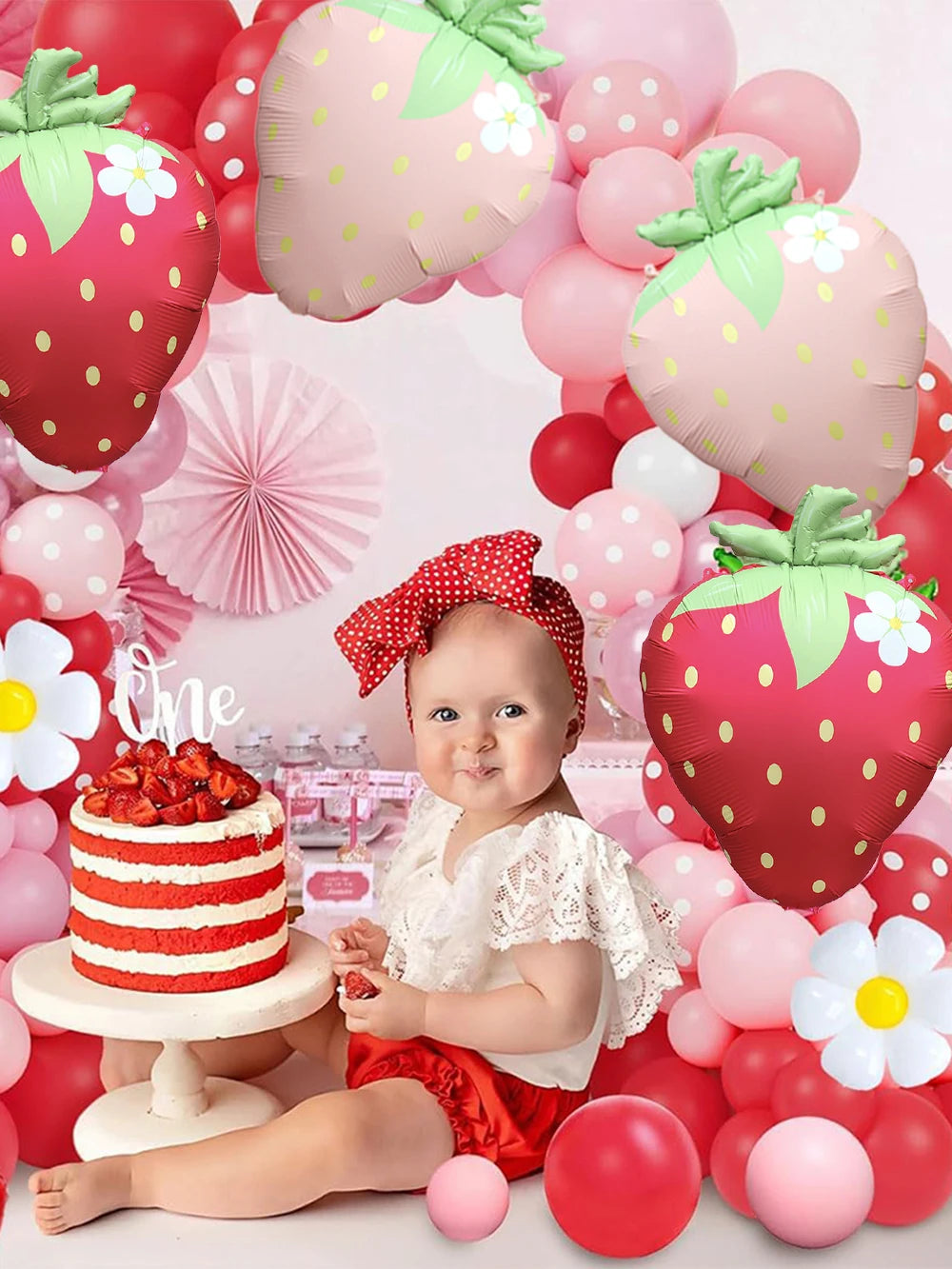 A baby is sitting in front of a large pink strawberry foil balloon surrounded by various pink and red balloons for a themed p