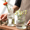 A hand holds a clear glass vase with a single orange rose and another small glass vase with yellow wildflowers on a wooden su