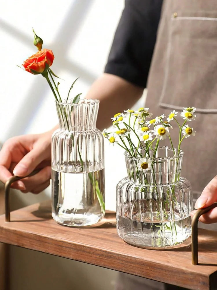 A hand holds a clear glass vase with a single orange rose and another small glass vase with yellow wildflowers on a wooden su
