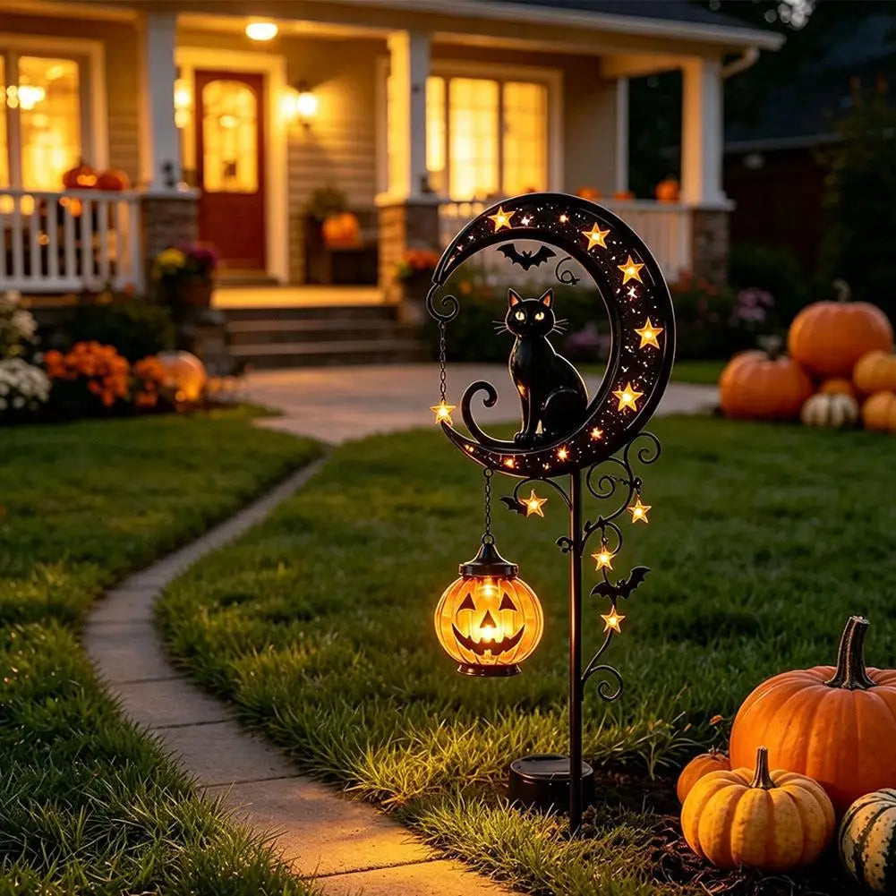 A black cat silhouette on a decorative metal moon with glowing lights, placed in a garden pathway surrounded by pumpkins.