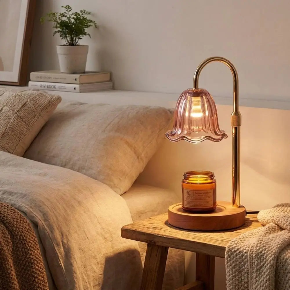 A brass adjustable height aromatherapy candle warmer lamp with a glass shade sits on a wooden table next to bedding.