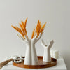 A white ceramic hand vase with golden wheat stalks is displayed on a wooden tray.