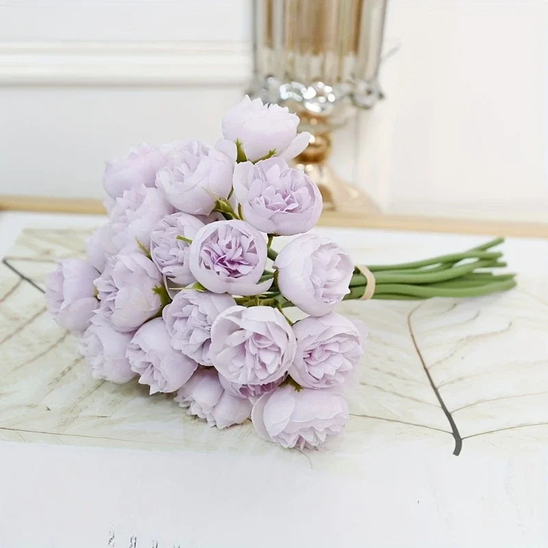 A bouquet of light purple silk peonies with green stems displayed on a white surface.