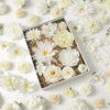 A collection of white and cream silk artificial flower heads arranged in a box with natural filler material.