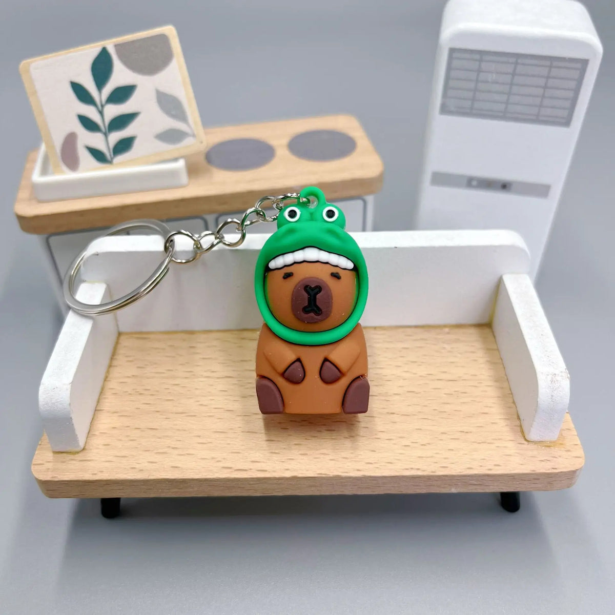 Pendentif de clé en forme de capybara en céramique avec peau verte et corps brun, posé sur un plateau en bois.