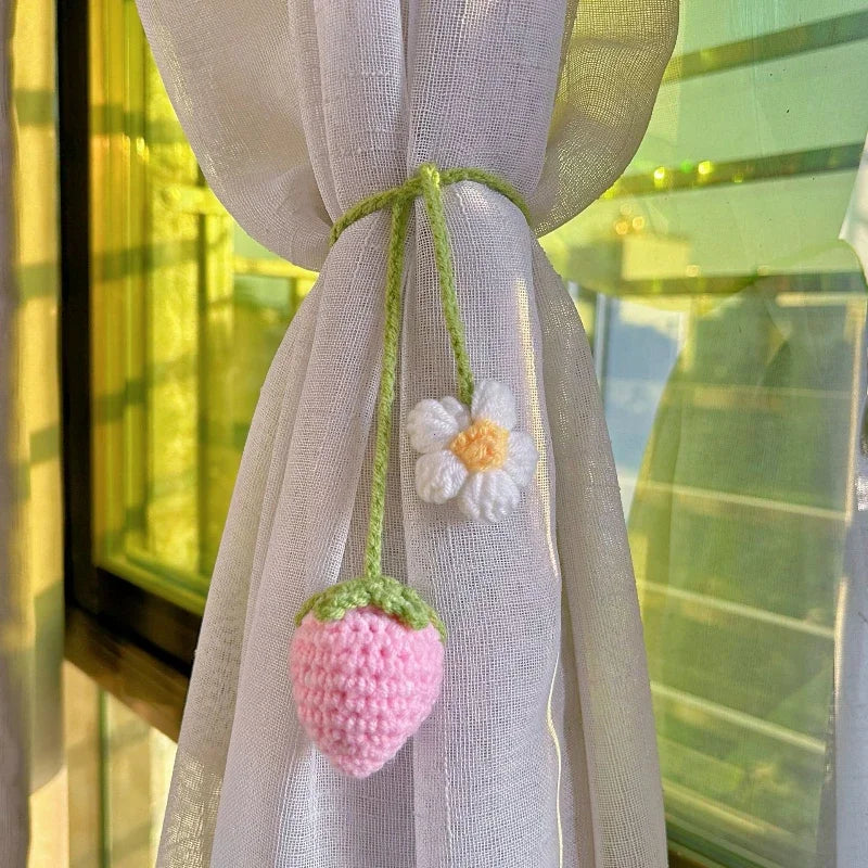 A hand-knitted hanging plant basket in pink with a white flower detail, tied with green twine, displayed near a window.