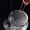 A hand is pouring water through a stainless steel colander into a glass bowl.