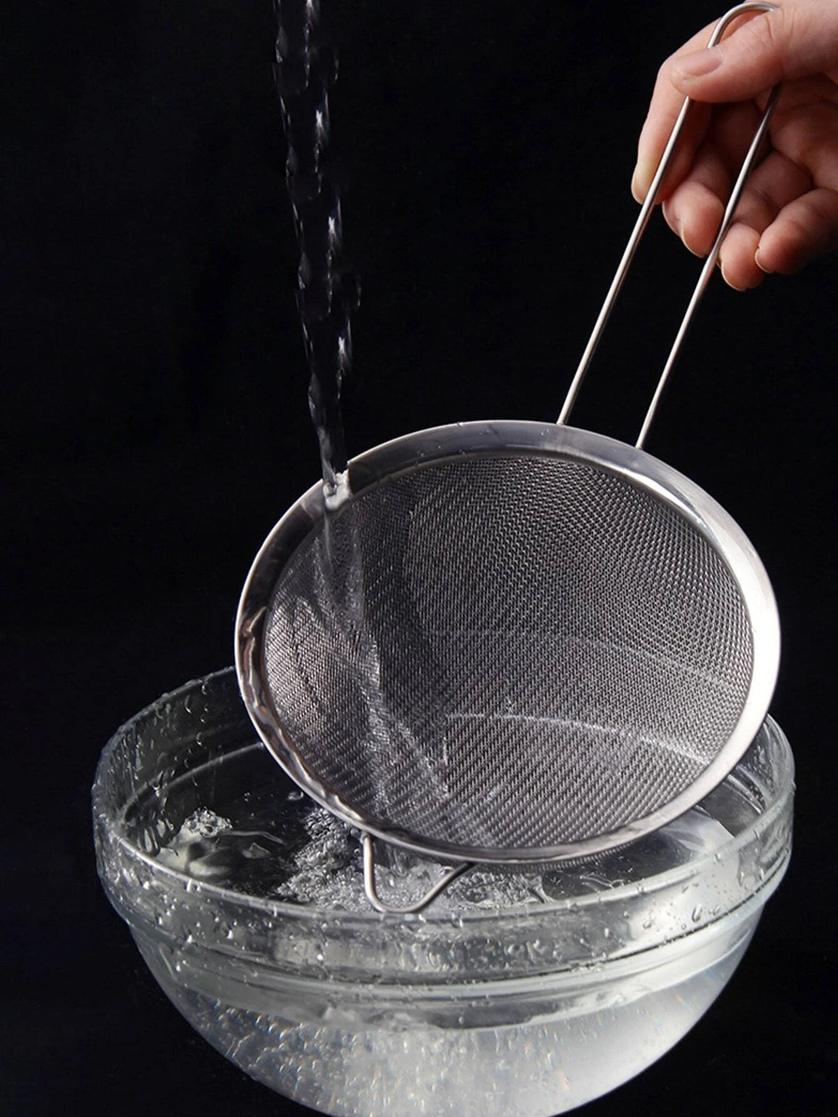 A hand is pouring water through a stainless steel colander into a glass bowl.