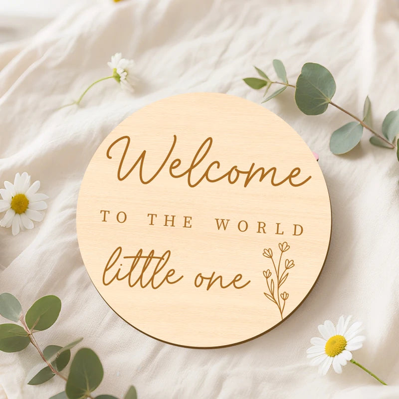 A round wooden sign with cursive text that reads Welcome to the world little one resting on a white fabric surrounded by gree