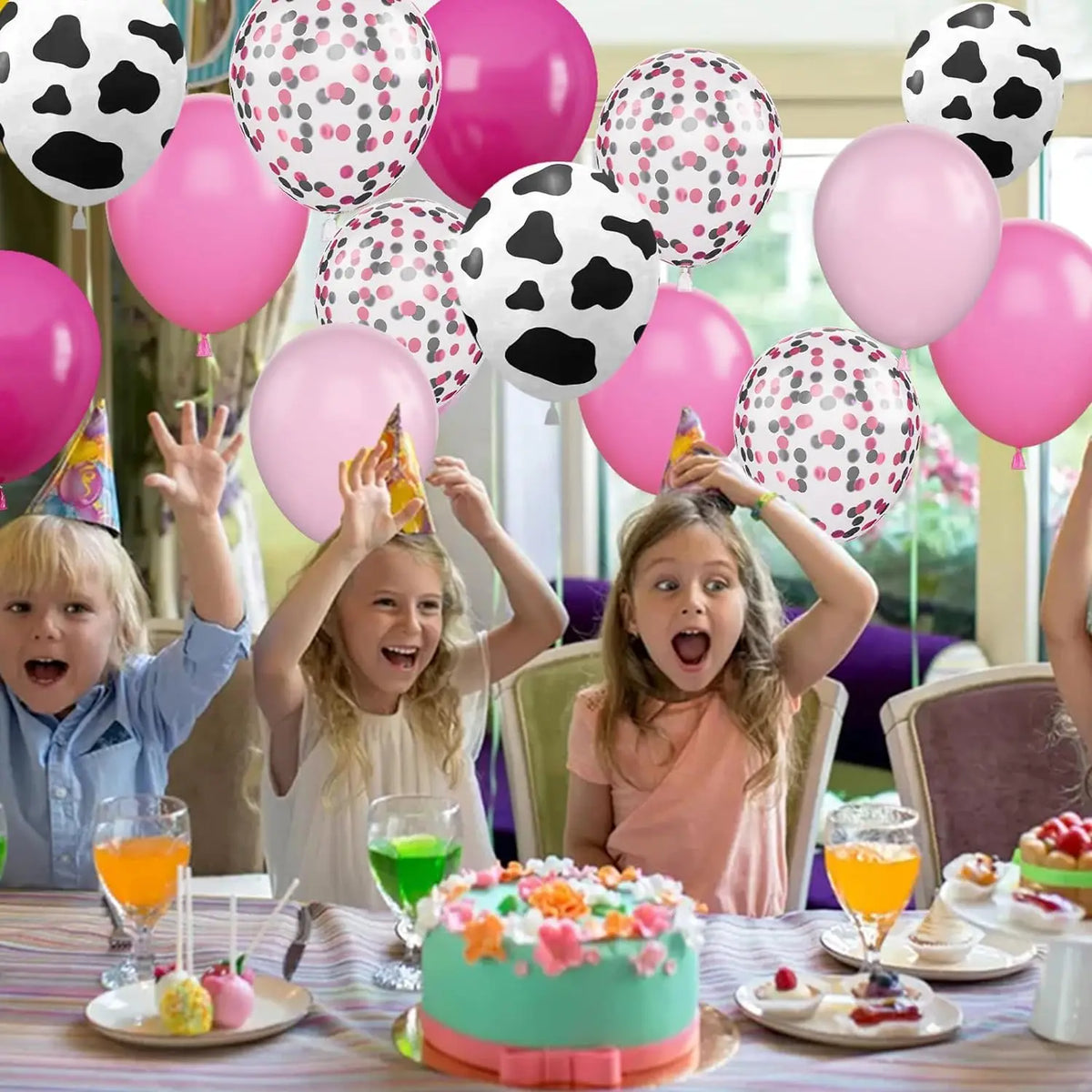 A table set with pink and white balloons, fruit, and a cake, surrounded by happy children.