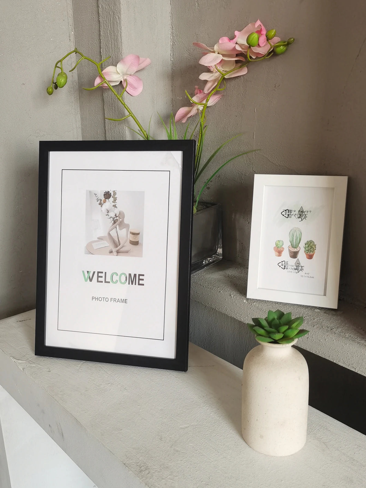 A black photo frame displaying the word WELCOME, resting on a light surface next to a potted pink orchid.