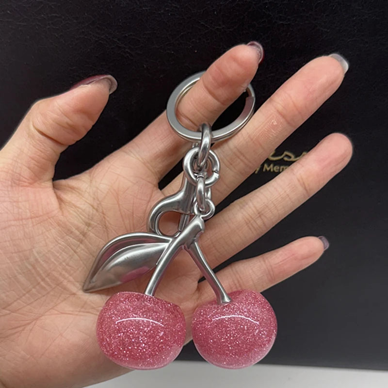 A silver charm featuring a pink glitter cherry pendant attached to a keychain, held by a hand.