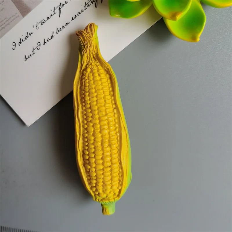 A bright yellow corn on the cob is placed next to a piece of paper with handwritten text and green leaves.