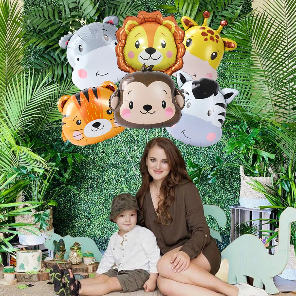 A woman sits with two young boys in front of a green foliage backdrop featuring foil balloons of safari animals like a lion, 