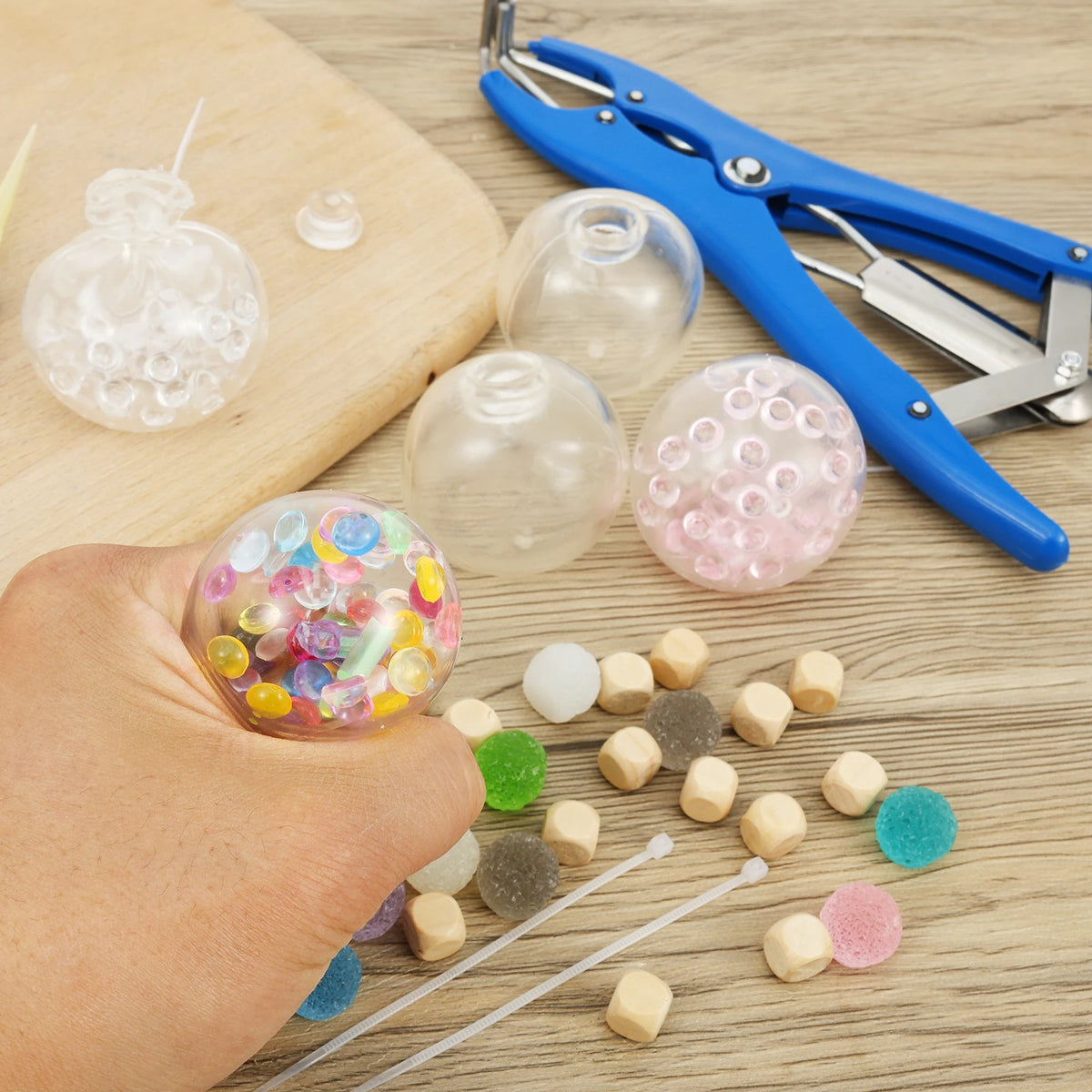 A hand holds a colorful stress ball, surrounded by various clear and colored plastic beads and pliers.