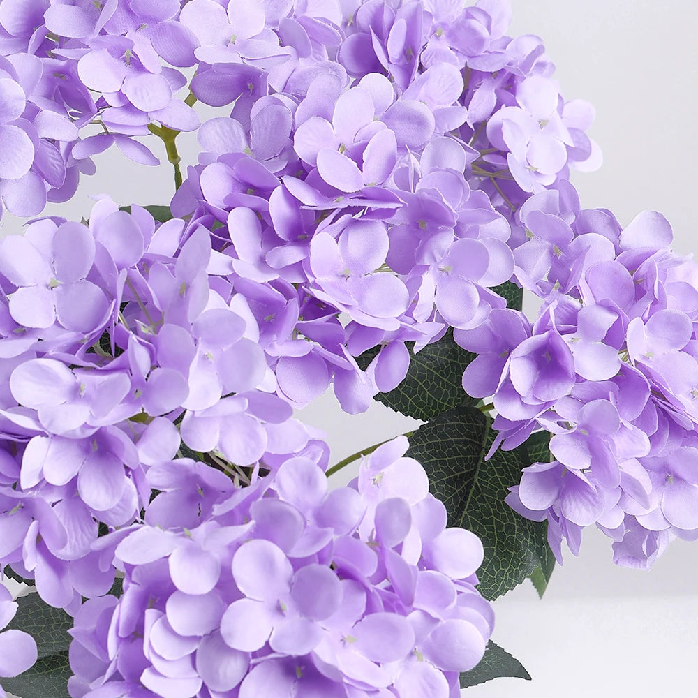 A close-up of a cluster of artificial purple hydrangea silk flowers on green leaves.