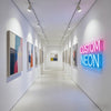 A brightly lit gallery hallway featuring framed artwork and a neon sign that reads CUSTOM NEON