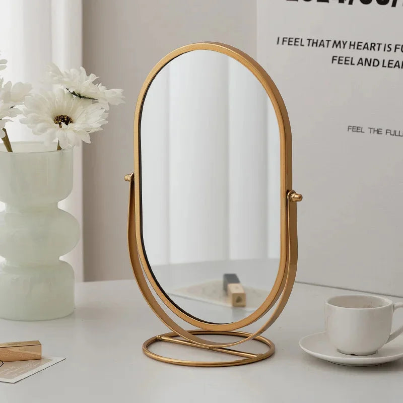 A gold-framed oval vanity mirror resting on a white surface next to a potted flower and a notebook.