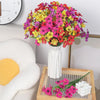 A vibrant bouquet of silk artificial flowers in shades of pink, yellow, and red arranged in a white vase on a wooden tray.