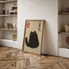 A framed canvas print featuring a black tiger cat illustration with Japanese text on a light wood background, displayed on a 