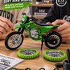 A green 1:12 scale dirt bike model is displayed on a wooden surface with spare tires and accessories.