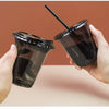 A hand holds a black disposable U-shaped coffee cup with a straw against a reddish-brown background.