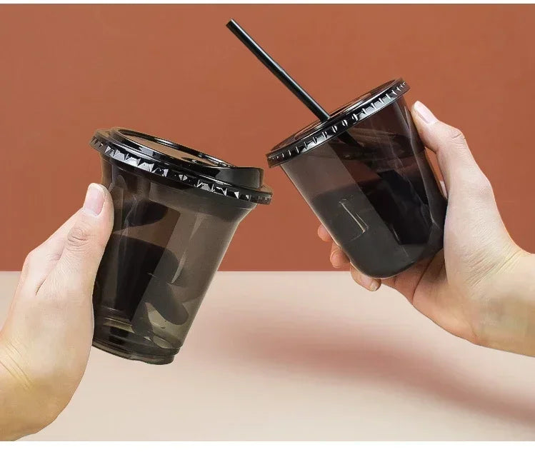 A hand holds a black disposable U-shaped coffee cup with a straw against a reddish-brown background.