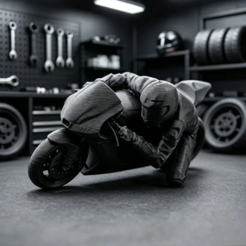 A dark figurine of a racing motorcycle rider in full gear is displayed on a desk with tools and tires in the background.
