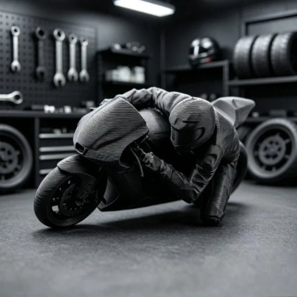 A dark figurine of a racing motorcycle rider in full gear is displayed on a desk with tools and tires in the background.