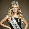 A blonde woman wearing a silver rhinestone birthday crown and a glitter sash against a gray background.