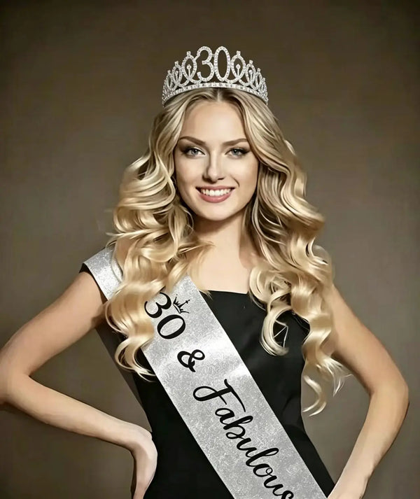A blonde woman wearing a silver rhinestone birthday crown and a glitter sash against a gray background.