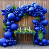 A royal blue balloon arch garland decorated with greenery and flowers on a wooden surface.