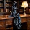 A black, hooded female figure figurine is displayed on a wooden desk next to a lamp in a library setting.