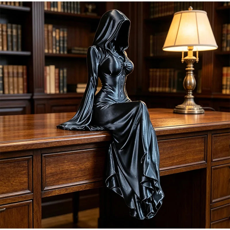 A black, hooded female figure figurine is displayed on a wooden desk next to a lamp in a library setting.