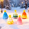 A collection of colorful, yellow and green resin Christmas tree charms resting in a snowy outdoor setting.