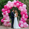 A bride and groom stand in front of a pink balloon arch decorated with various shades of pink balloons.
