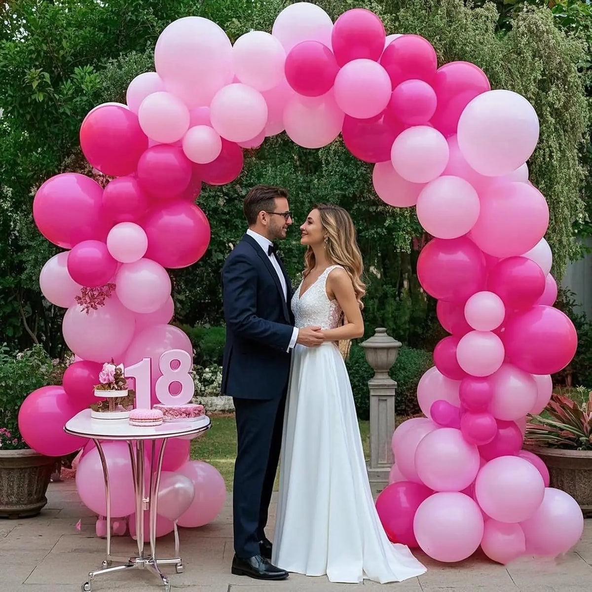 A bride and groom stand in front of a pink balloon arch decorated with various shades of pink balloons.