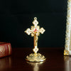 A golden metal Jesus cross ornament with a decorative base, displayed on a dark wooden surface.
