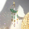 A gold moon-shaped crystal suncatcher with green crystals and a rainbow prism hanging against a bright background.