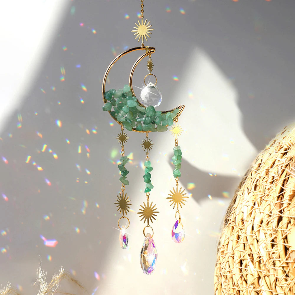 A gold moon-shaped crystal suncatcher with green crystals and a rainbow prism hanging against a bright background.