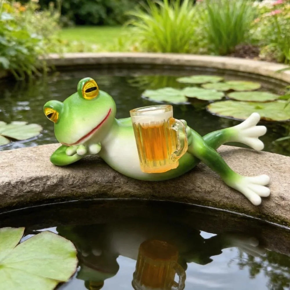 Figurita de rana de resina verde sosteniendo un vaso, reposando sobre una piedra junto a plantas de agua.