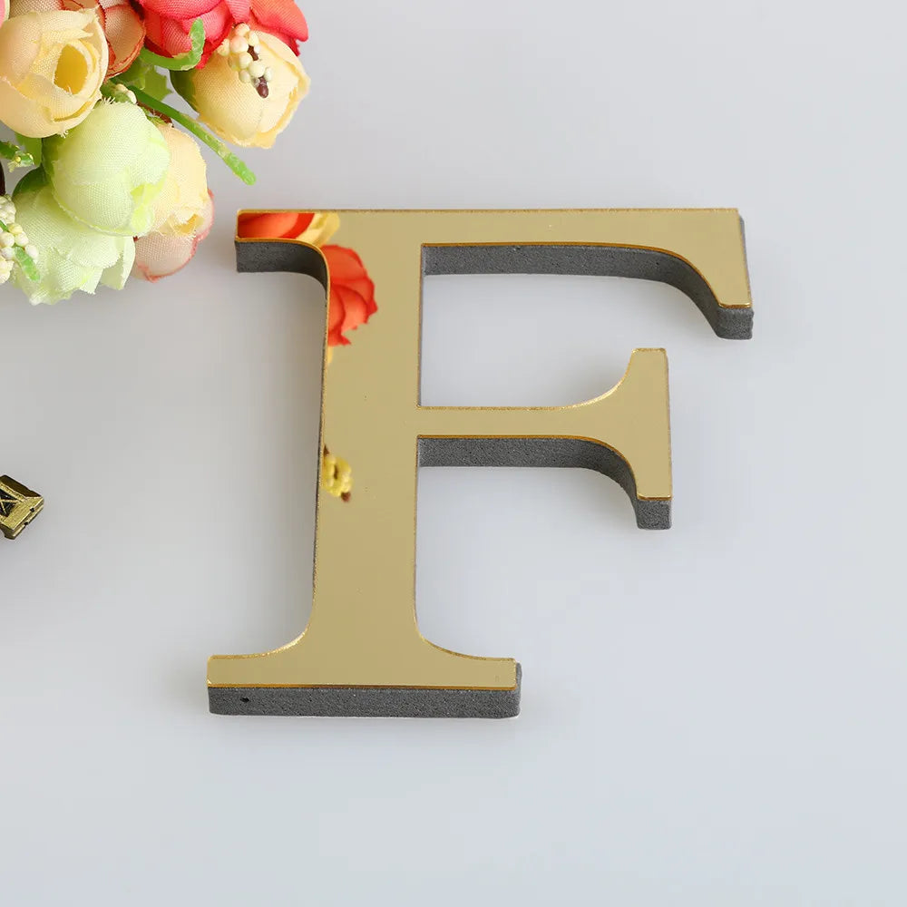 A gold letter F cutout for a mirror wall decal set, placed on a white surface with flowers in the background.