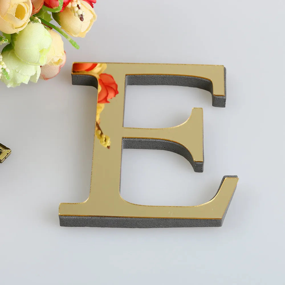 A gold letter E cutout for a mirror wall decal set, placed next to some light-colored flowers.