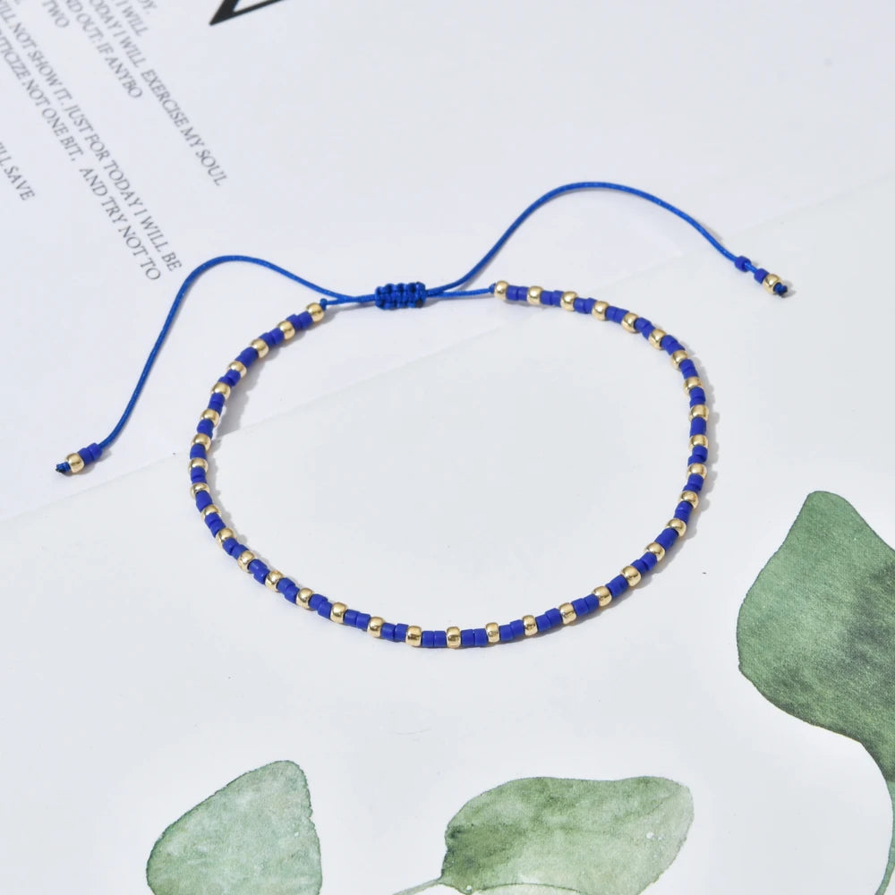 A handmade boho glass bead bracelet with an adjustable cord, featuring blue and gold beads, laid on white paper with green wa