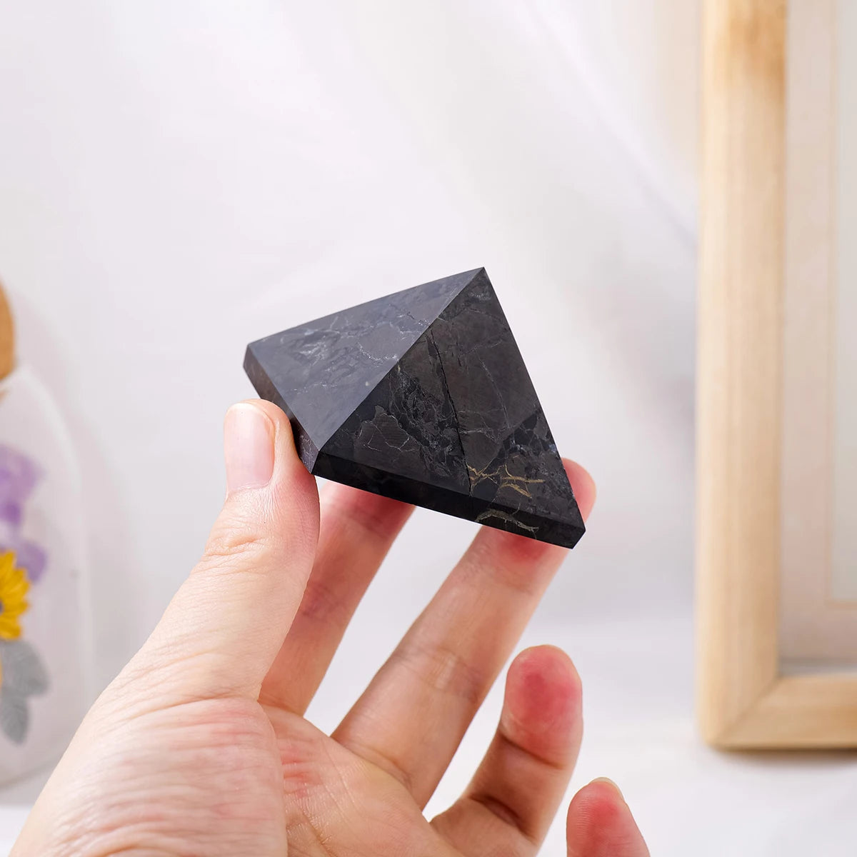 Una mano sostiene una pirámide de cristal de shungita negra, mostrando sus facetas sobre un fondo blanco.