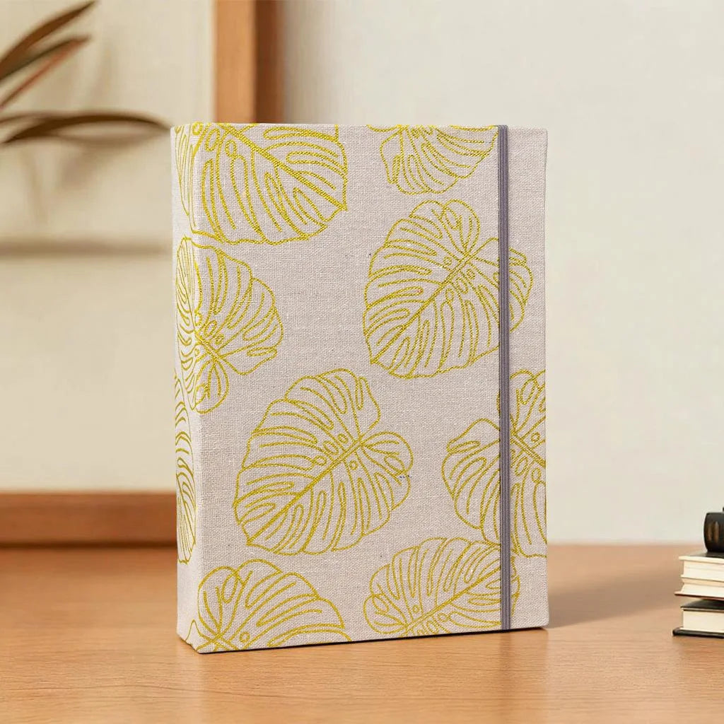 Helles beige Fotoalbum mit einem Cover aus gelben Linienzeichnungen von Monstera-Blättern.