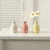 Three ceramic mini vases in white, pink, and yellow holding dried flower stems on a white tabletop.