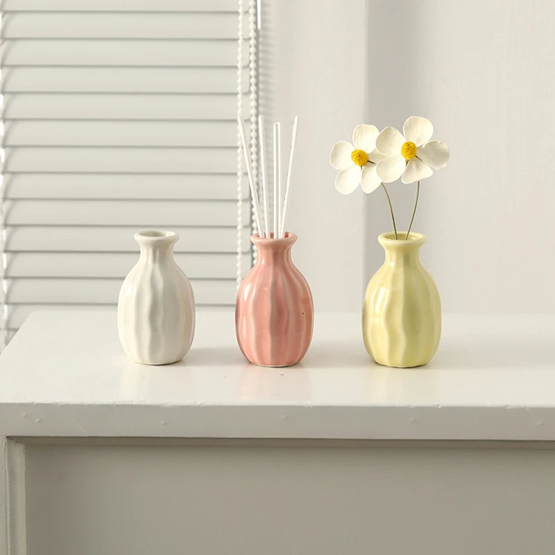 Three ceramic mini vases in white, pink, and yellow holding dried flower stems on a white tabletop.