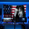 A man with long dark hair and a beard holds a banner featuring the text TOP 10 MUST-SEE NORMAN REEDUS on a patriotic backgrou