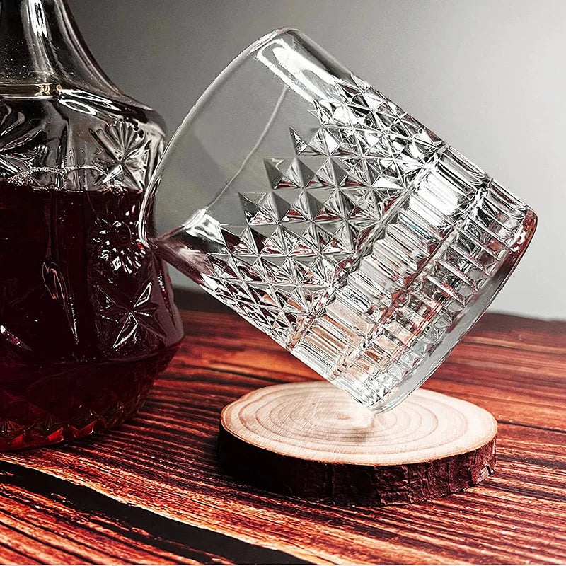 A clear whiskey glass with a diamond-cut pattern rests on a wooden slice next to a dark bottle.